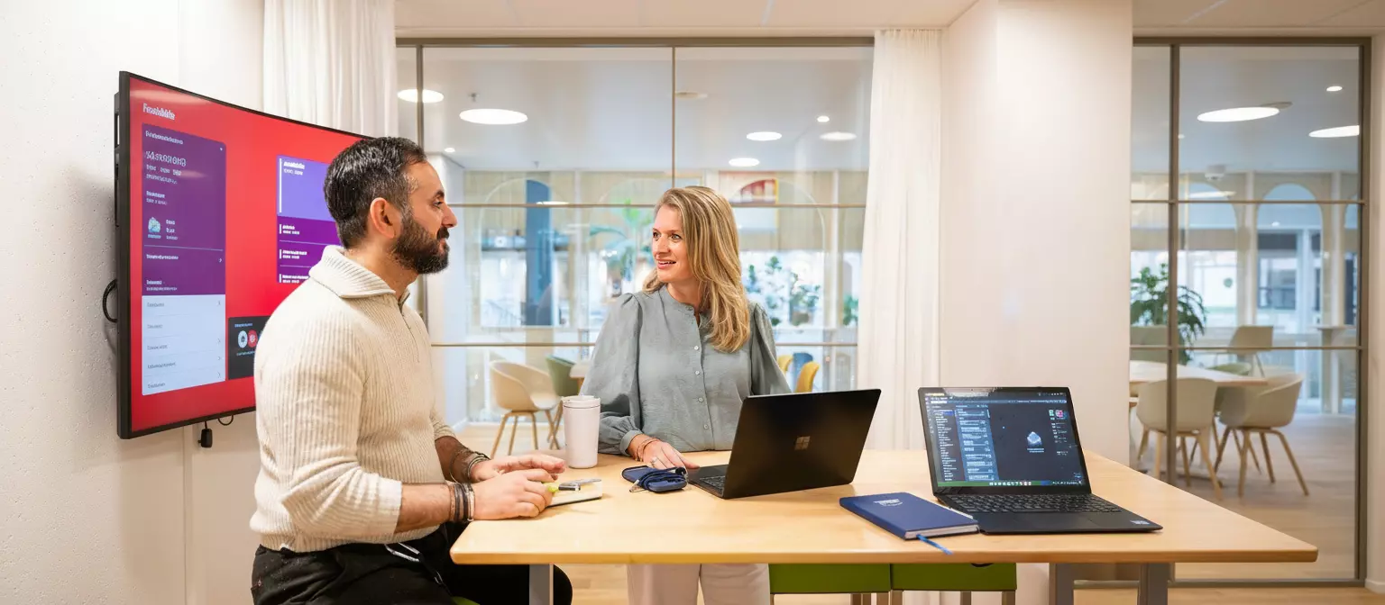 Twee collega's zitten met hun laptops op banken in overleg op kantoor.