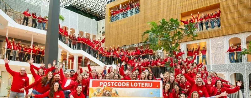 Grote groep collega's met rode Postcode Loterij-jasjes staan met een grote cheque met de totale jaaropbrengst te juichen op kantoor.