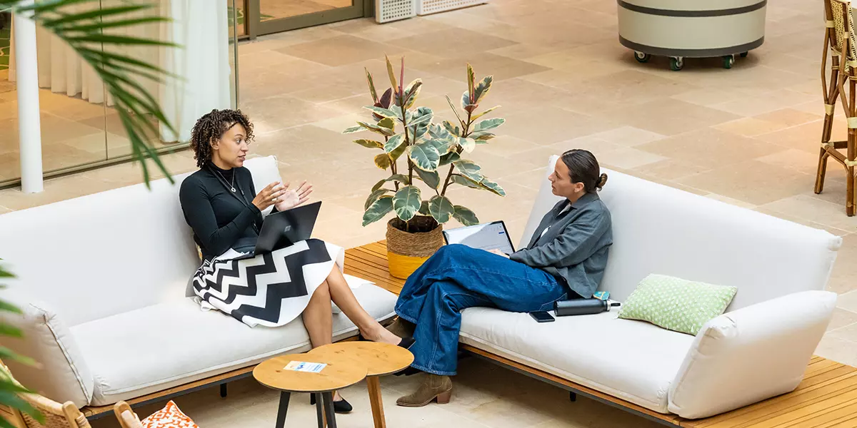 Twee collega's zitten met hun laptops op banken in overleg op kantoor.